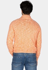 Oranje gebreide trui met een gestructureerde afwerking, voorzien van een hoge kraag. Gecombineerd met blauwe spijkerbroek, waarbij de achterkant en geribde manchetten zichtbaar zijn.
