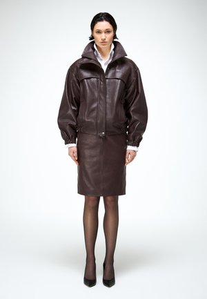 Vrouw in donkerbruine leren jas en bijpassende rok, met doorschijnende zwarte panty's en zwarte hoge hakken, staand tegen een witte achtergrond.