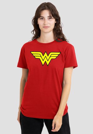 Young woman with long brown hair wearing a red T-shirt with a large yellow Wonder Woman logo on the chest, standing against a plain background.