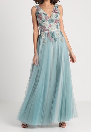 Frau trägt ein bodenlanges, hellblaues Tüllkleid mit floraler Stickerei am Oberteil und einem tiefen V-Ausschnitt.