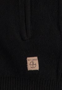 Sudadera de forro polar negra con una cremallera corta, que presenta un acabado texturizado y una etiqueta de tela marrón que dice "Key Largo" cosida en la parte inferior.