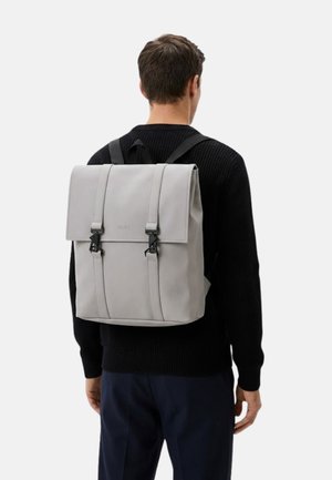 Mochila gris hecha de material liso con una solapa, doble hebilla y costuras verticales. Se ven correas ajustables para los hombros.