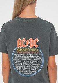 Szara bawełniana koszulka z logo AC/DC w kolorze czerwonym i pomarańczowym, z niebieskim okrągłym tekstem zawierającym listę miejsc trasy koncertowej "Highway to Hell" z 1979 roku.