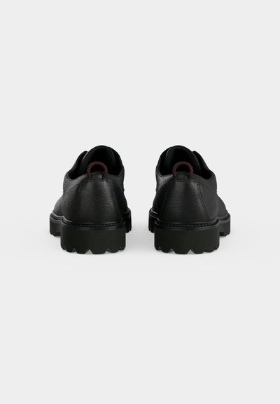 Paire de chaussures en cuir noir avec semelles en caoutchouc épaisses et languettes rouges et noires, vues de dos sur un fond uni.