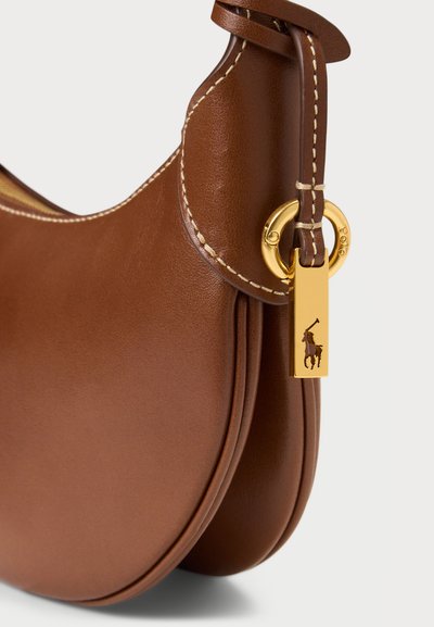 Ruda odinė rankinė, turinti kreivinę formą, matomą siuvimą, auksinį metalinį akcentą ir kabinamą žavesį su logotipo detale.