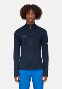 Marineblauwe jas met een geaccentueerde rits aan de voorkant, verticale textuurpanelen, hoge kraag en een oranje ritsaccent. Voorzien van het MAMMUT-logo.