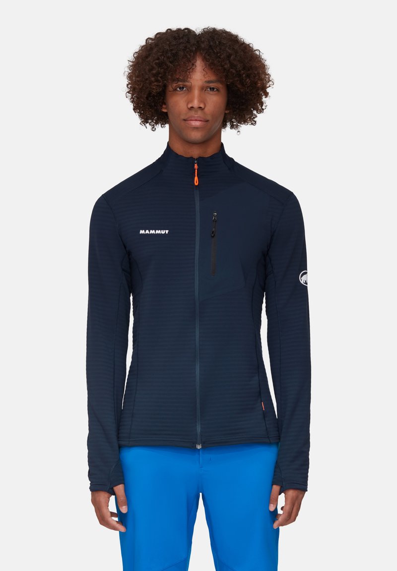 Marineblauwe jas met een geaccentueerde rits aan de voorkant, verticale textuurpanelen, hoge kraag en een oranje ritsaccent. Voorzien van het MAMMUT-logo.