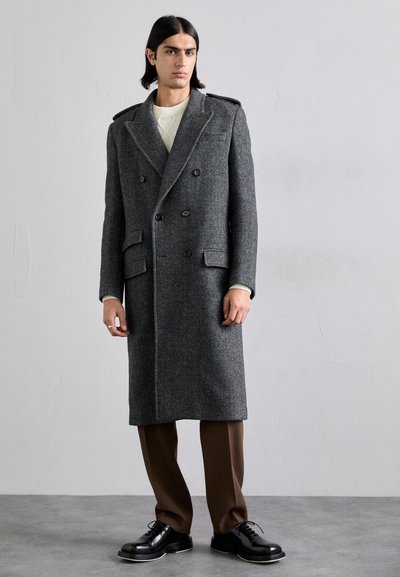 Grijze wolmix dubbele breasted jas met schouderepauletten, klepzakken en contrasterende stiksels, gecombineerd met bruine broek en zwarte schoenen.