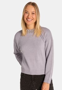 Maglione viola chiaro con un motivo testurizzato vicino al colletto, dotato di polsini e orlo a coste. Indossato sopra pantaloni neri con pois bianchi.