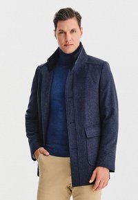 Morska wełniana płaszcz w kolorze granatowym z kołnierzem, przednimi kieszeniami i zapięciem na guziki, noszony na ciemnoniebieskim golfie i beżowych spodniach.