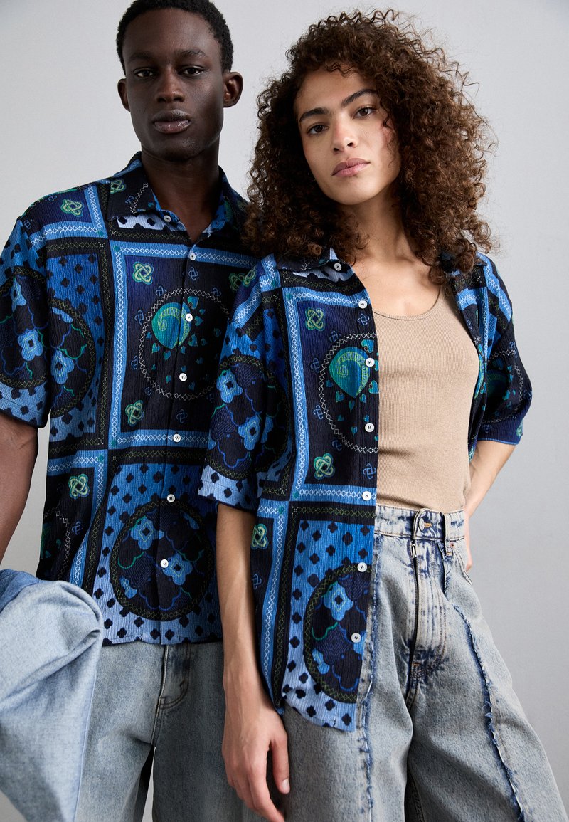 Les chemises à manches courtes présentent un motif bleu et noir avec des motifs floraux, associées à un jean en denim clair et un débardeur beige.