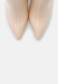 Tacones en charol beige con punta afilada, superficie lisa, detalles de costura, silueta esbelta y sin herrajes visibles.