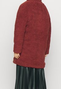 Burgundy fleecejacka med en mjuk, texturerad yta, hög krage och avslappnad passform, kombinerad med en svart veckad kjol.