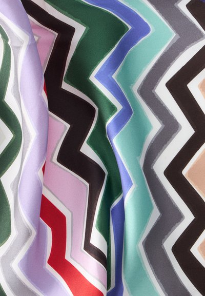 Tissu avec motif zigzag coloré en vert, noir, rouge, violet, bleu, sarcelle, gris et beige, montrant des plis et une texture.