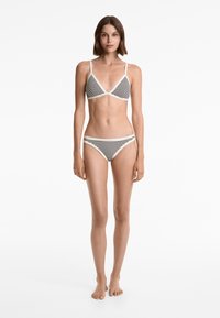 Ung kvinna som står barfota iförd en svartvit randig bikini med tunna remmar mot en vit bakgrund.