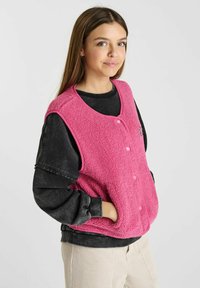 Roze fleece mouwloze vest met een ronde hals, voor knopen en zijzakken, gedragen over een zwarte longsleeve shirt.