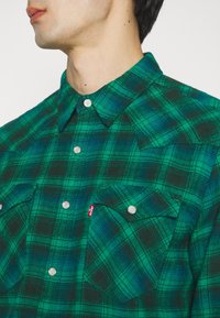 Camisa a cuadros en verde y negro con collar puntiagudo, botones de presión plateados, dos bolsillos en el pecho y una pequeña etiqueta con logo rojo. Textura de tela suave.