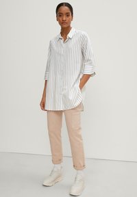 Camicia a righe bianca e beige, abbinata a pantaloni cargo beige chiaro e sneakers bianche voluminose. La camicia ha maniche arrotolabili.