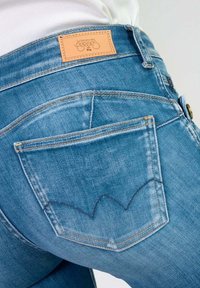 Gros plan sur la poche arrière d'un jean en denim bleu avec un détail courbé cousu et une étiquette de marque en cuir beige sur la ceinture.