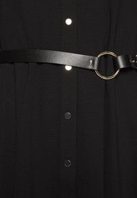 Robe noire avec un devant boutonné et une taille cintrée par une ceinture en cuir noir, agrémentée d'une boucle circulaire dorée et d'éléments en argent.