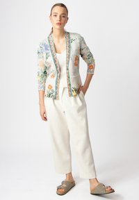 Cardigan a fantasia floreale in tessuto leggero, con fiori verdi, arancioni e viola; abbinato a una canotta bianca e a pantaloni color crema.