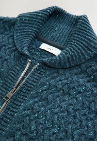 Tumši zils adīts džemperis ar augstu apkakli, kuram ir teksturēts vadiņu raksts un rāvējslēdzējs, akcentēts ar terakotu punktiem.