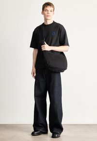 Μαύρο φαρδύ t-shirt με στρογγυλή κεντημένη ετικέτα, σε συνδυασμό με φαρδιά σκούρα τζιν και μαύρη υφασμάτινη τσάντα ώμου. Ομαλές υφές.