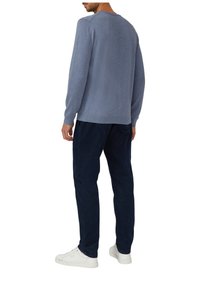 Mann in einem hellblauen Pullover, dunklen Marinehosen und weißen Sneakers, der mit dem Rücken zu einem weißen Hintergrund steht.