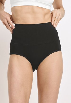 Frau trägt hoch taillierte schwarze Unterwäsche und ein weißes, kurz geschnittenes Oberteil, die Hände justieren den Bund vor einem einfarbigen hellen Hintergrund.