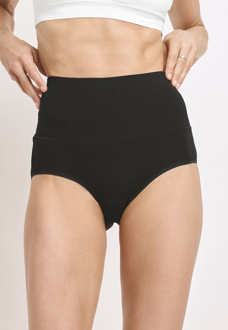 Kobieta w czarnych majtkach o wysokim stanie i białym krótkim topie, ręce poprawiające pasek na tle jasnego, jednolitego tła.