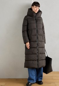 Lang, donkerbruine pufferjas met een hoge kraag, voorzien van een ritssluiting aan de voorkant, grote zakken en een gestructureerde afwerking. Gecombineerd met brede blauwe jeans.