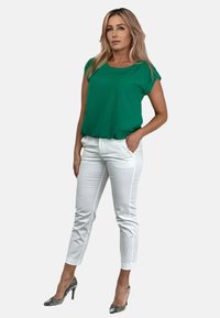 Zielony krótki top z luźnym krojem, w zestawieniu z dopasowanymi białymi spodniami i wzorzystymi butami na obcasie, na gładkim tle.