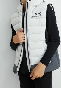 Osoba nosząca białą i szaro-srebrną kamizelkę puchow�ą z napisem "NYC _STAY READY" na czarnej koszulce z długim rękawem, zapinająca kamizelkę.