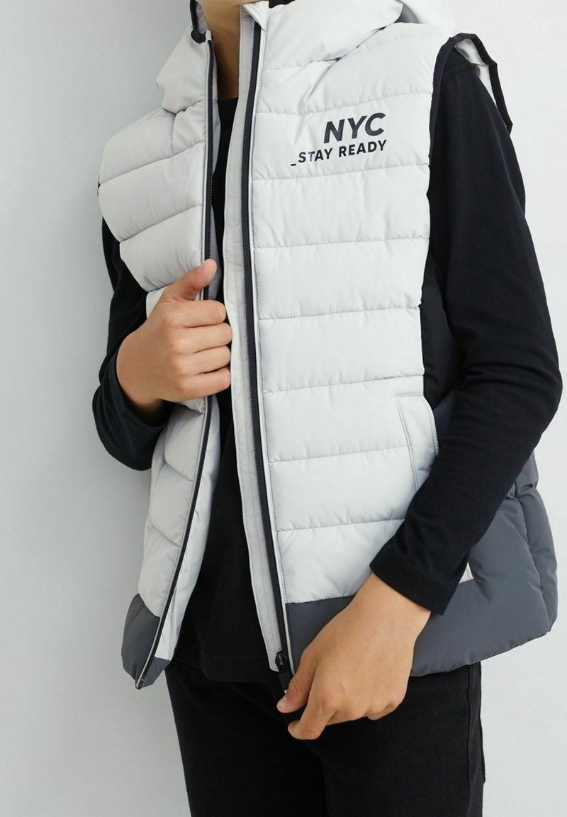 Osoba nosząca białą i szaro-srebrną kamizelkę puchową z napisem "NYC _STAY READY" na czarnej koszulce z długim rękawem, zapinająca kamizelkę.