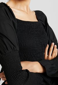 Svart smockad topp med fyrkantig halsringning, puffade ärmar och strukturerad detaljering på ärmar och överdel. Slät tygfinish.