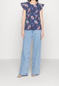 Bluse mit floralem Muster und kurzen Rüschenärmeln in Marineblau, kombiniert mit hellblauen, weiten Jeans, die einen entspannten Schnitt und eine gerade Silhouette aufweisen.