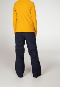 Žlutý fleecový top s dlouhým rukávem v kombinaci s námořnickými lyžařskými kalhotami, které se vyznačují volným střihem a odolnou látkou. Kalhoty mají rovný střih a kapsy.