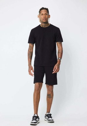 Homme avec des tatouages et des dreadlocks courts debout face à l'avant, portant un t-shirt noir, un short noir et des baskets noires et blanches.