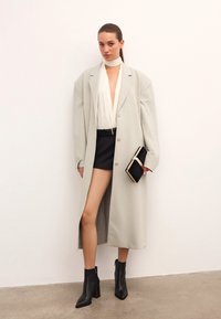 Šviesiai pilka ilga švarko striukė ant baltos plisėtės bluzės ir juodų šortų. Papuošta juodomis kulkšninių botais ir mažu juodu rankiniu krepšiu su auksinėmis detalėmis.