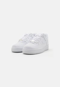 White sneakers featuring a textured upper, round toe, flat laces, and a rubber sole with a patterned tread. Distinctive logo detail present.