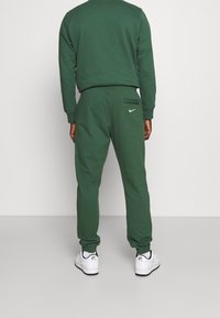 Grön bomullströja och joggingbyxor med elastiska muddar, som har en liten vit Nike-logga på bakfickan och klassiska vita sneakers.