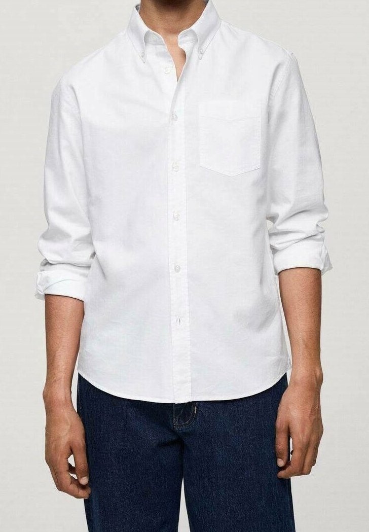 Homme portant une chemise blanche à manches longues boutonnée avec les manches retroussées et un jean bleu foncé sur un fond uni.