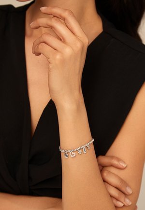 Primer plano de una muñeca que lleva una pulsera de dijes plateada con corazones y dijes rectangulares, la mano descansando cerca del cuello, se ve una camiseta negra sin mangas.