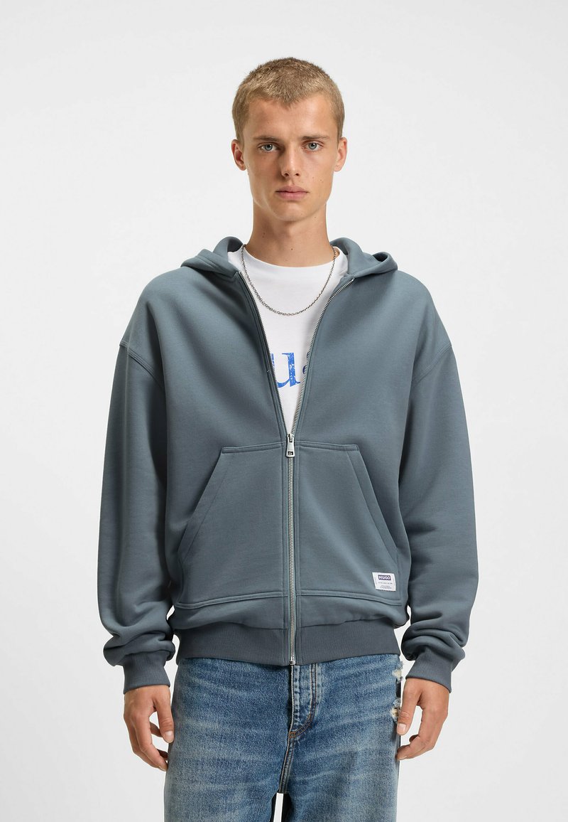 Grijze zip-up hoodie met een voorzak, voorzien van een trekkoordkap en ribgebreide manchetten, gedragen over een wit t-shirt. Gestructureerde stof.