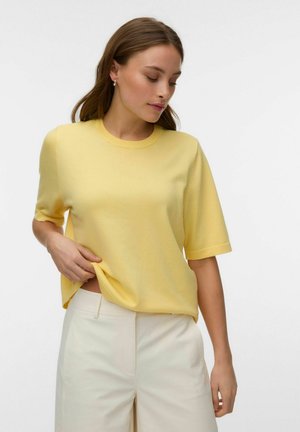 Femme portant un pull jaune à manches courtes et un pantalon blanc taille haute, tenant l'ourlet du pull tout en regardant vers le bas.