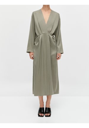 Woman wearing a long, loose, olive-green dress with a deep V-neck and long sleeves, paired with black open-toe mule shoes.