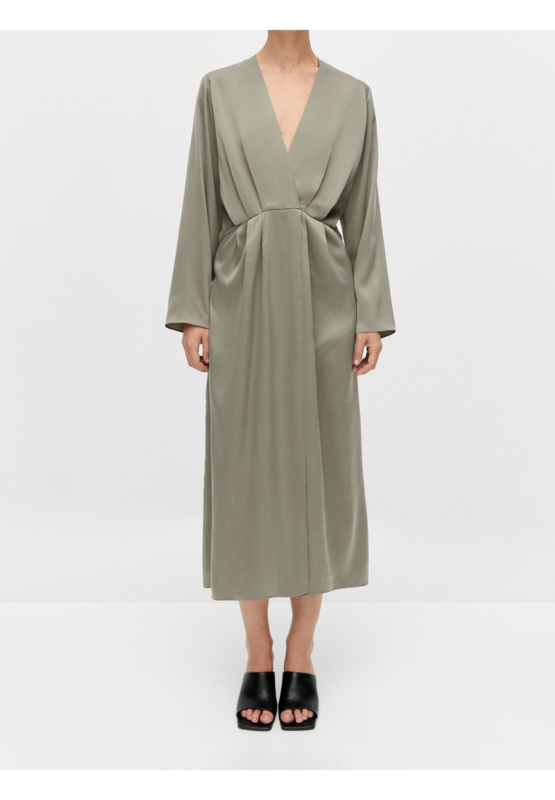 Femme portant une longue robe ample olive avec un décolleté en V profond et des manches longues, associée à des mules noires à bout ouvert.