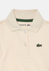 Kremičasti Lacoste polo majica s dva bijela gumba, zelenim rubom unutar ovratnika i zelenim krokodilom na prsima.