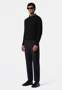 Pull noir, chemise à col gris, pantalon bleu marine et baskets noires. Le modèle porte des lunettes de soleil sombres, se tenant contre un fond uni.