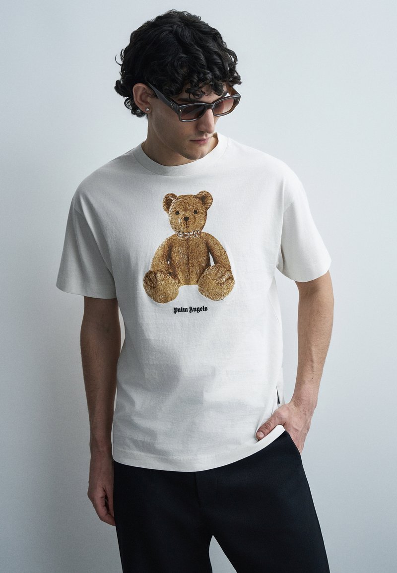 Homme portant des lunettes de soleil et un t-shirt blanc avec un grand motif d'ours en peluche et le texte "Palm Angels", debout devant un fond uni.